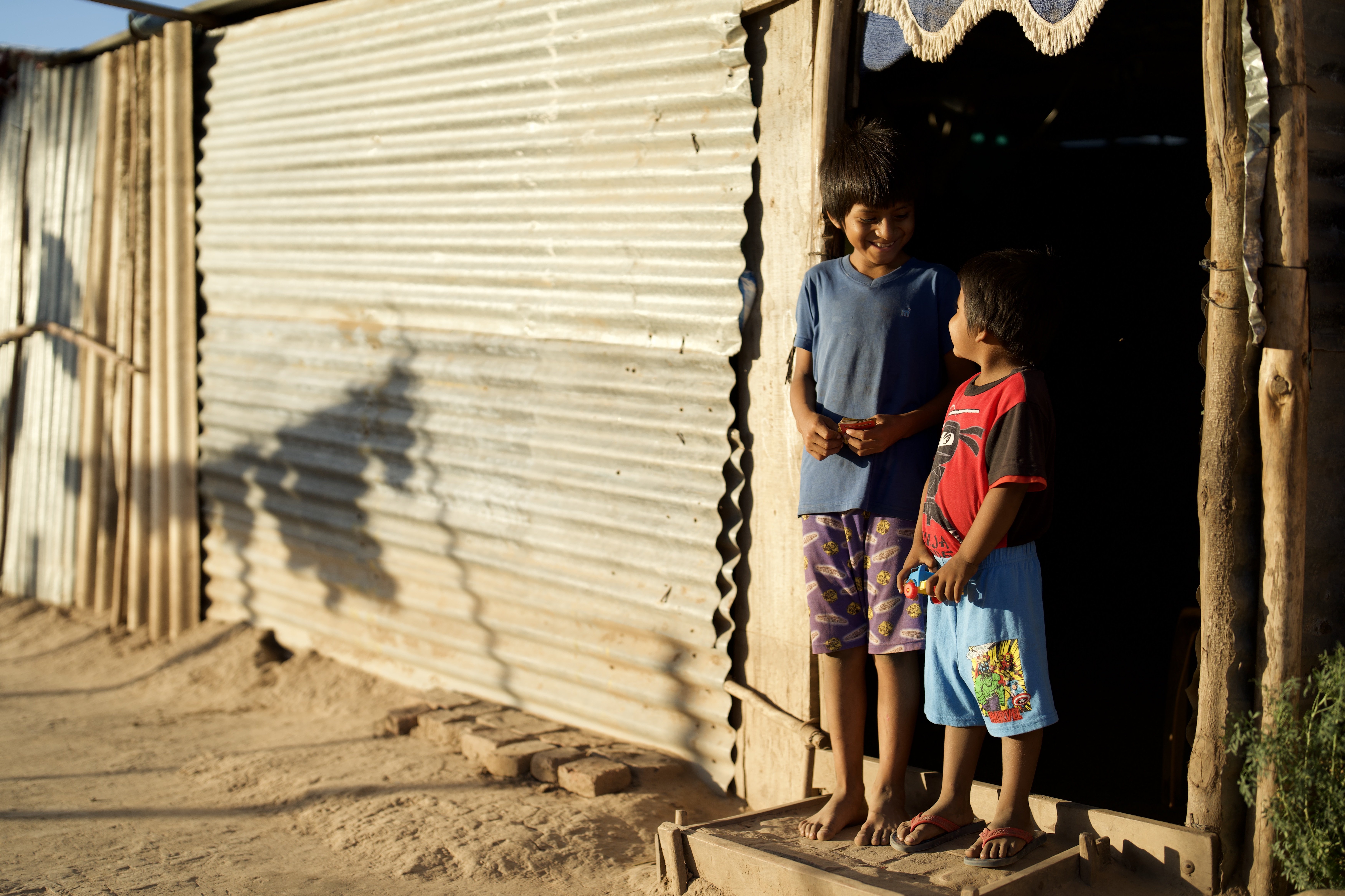 Two young boys stand outside a home made from metal sheets and wood and smile at one another.